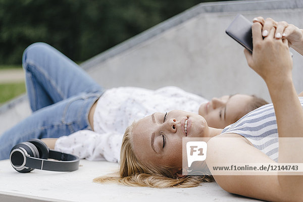 Zwei junge Frauen mit Handy und Kopfhörer auf einer Rampe im Skatepark liegend