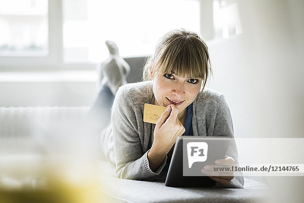 Lächelnde Frau auf der Couch liegend mit Kreditkarte und Tablette