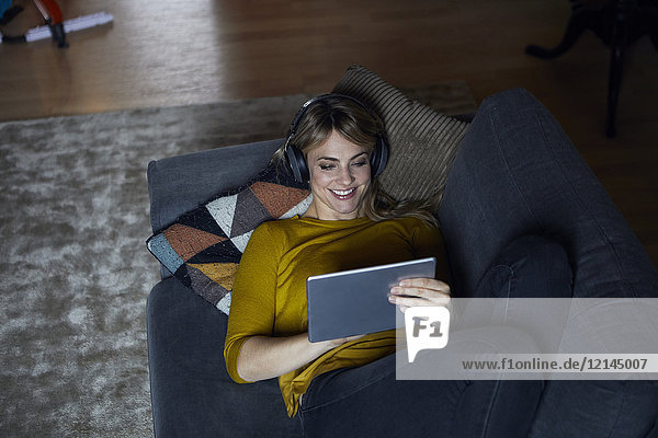 Porträt einer lächelnden Frau  die mit Kopfhörer und Tablett auf der Couch liegt.