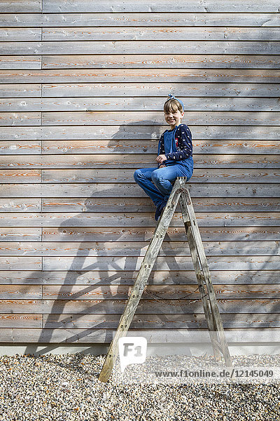 Lachendes Mädchen auf einer Leiter vor einer Holzfassade sitzend