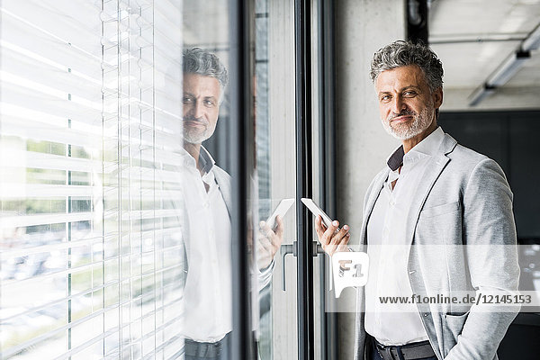 Porträt des lächelnden reifen Geschäftsmannes am Fenster im Büro mit Handy