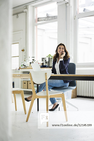 Porträt einer lachenden Geschäftsfrau am Telefon  die am Schreibtisch in einem Loft sitzt.