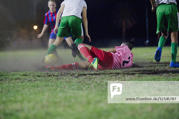 Young female soccer player falling  kicking the ball playing soccer on field at night