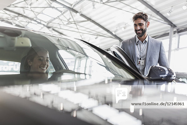 Lächelnder Autoverkäufer und Kundin auf dem Fahrersitz des Neuwagens im Autohaus-Showroom