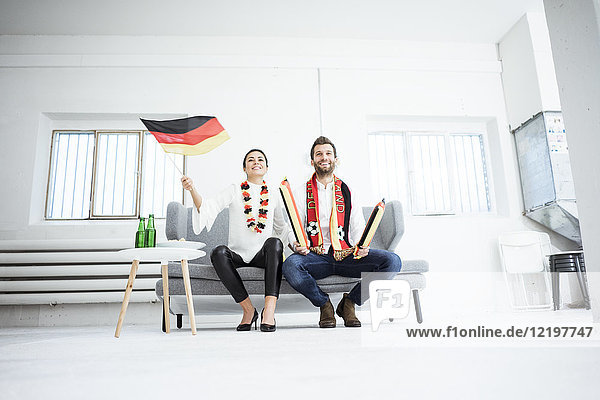 Fröhliches deutsches Fußballfanpaar sitzt auf der Couch und schaut fern.