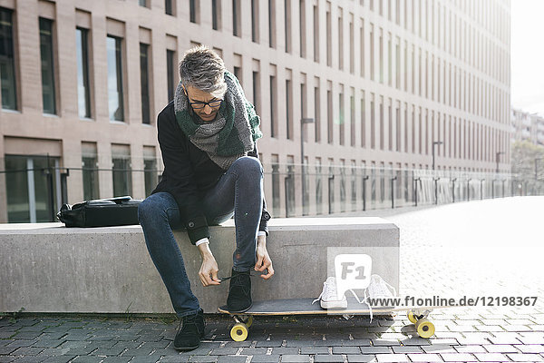 Freelancer with longboard sitting on bench tying his shoes