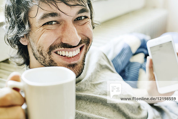 Porträt eines lachenden jungen Mannes auf dem Bett liegend mit Smartphone und Tasse Kaffee