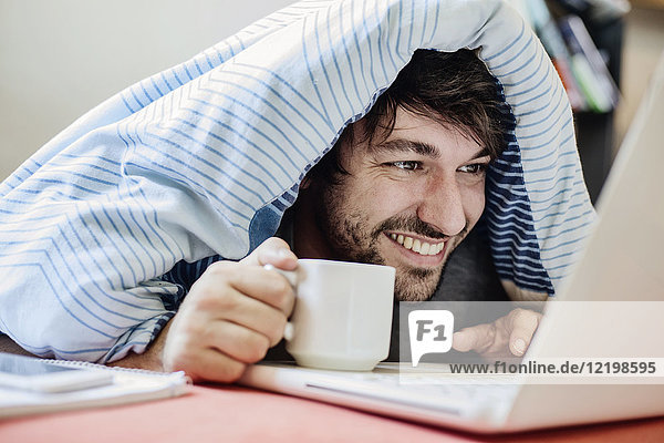Porträt eines lachenden jungen Mannes auf dem Bett liegend mit einer Tasse Kaffee mit Laptop