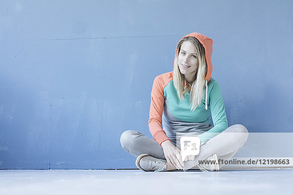 Porträt einer lächelnden Frau in Kapuzenjacke auf dem Boden vor der blauen Wand sitzend