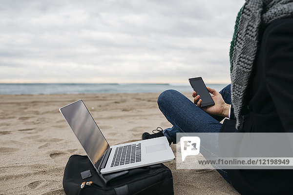 Businessman sitting on the beach in winter using laptop and cell phone  partial view