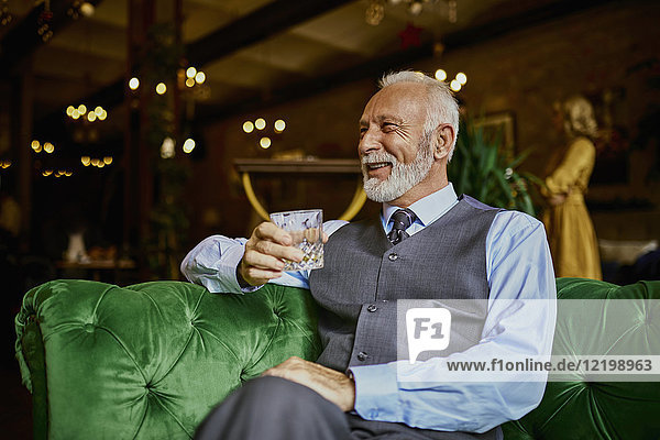 Portrait of elegant senior man sitting on couch in a bar holding tumbler