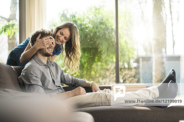 Smiling woman covering her boyfriend's eyes on sofa at home