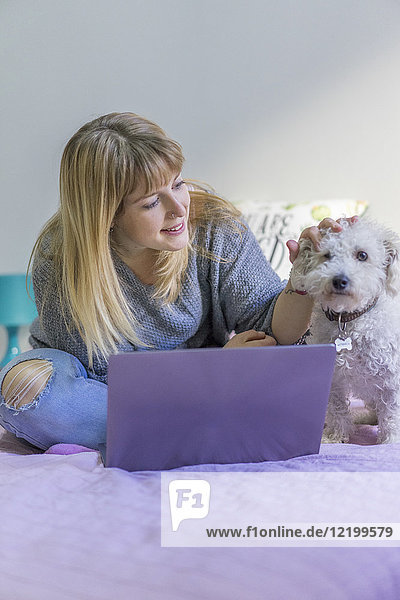 Frau mit Laptop sitzt auf dem Bett und streichelt ihren Hund.