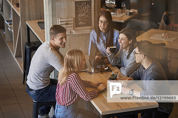Group of happy friends sitting together in a cafe with laptop and drinks