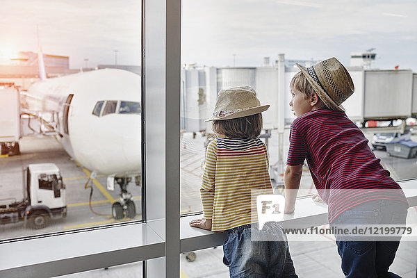 Zwei Kinder mit Strohhut schauen durchs Fenster zum Flugzeug auf dem Vorfeld