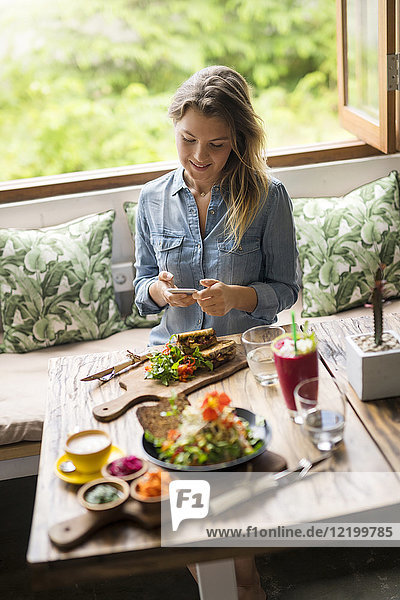 Frau fotografiert Essen mit Smartphone im gemütlichen Café vor dem Fenster