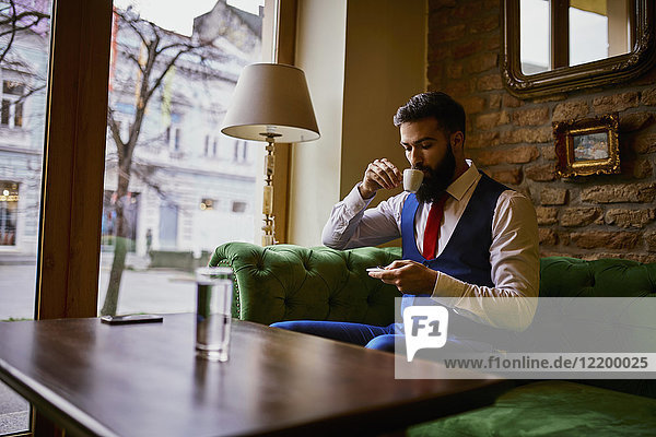 Modischer junger Mann sitzt auf der Couch in einem Café  trinkt Kaffee und benutzt das Handy.