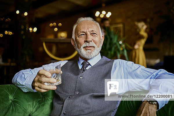 Porträt eines eleganten älteren Mannes  der auf einer Couch in einer Bar mit Becher sitzt.