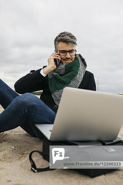 Portrait of businessman on the phone sitting on the beach in winter using laptop