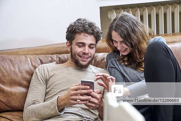 Junger Mann und junge Frau sitzen auf der Couch und teilen sich Handy und Kopfhörer.