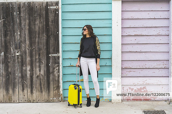 Junge Frau mit gelber Trolley-Tasche vor Holzfassade