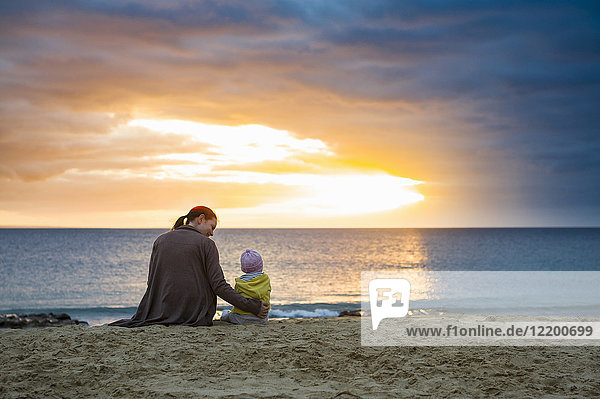 Mother with little daughter sitting on the beach at sunset