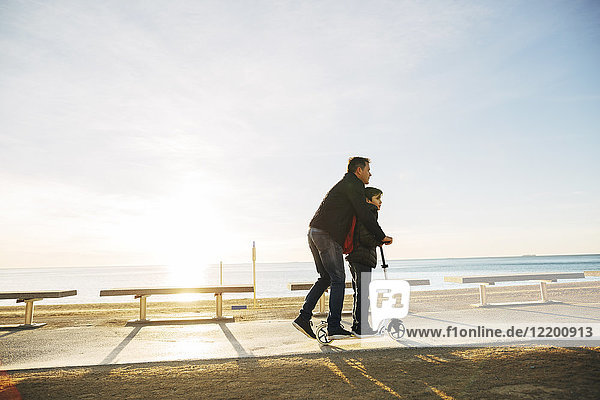 Vater und Sohn fahren Roller auf der Strandpromenade bei Sonnenuntergang