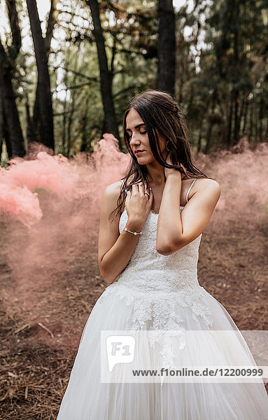 Frau mit geschlossenen Augen trägt ein Hochzeitskleid im Wald  umgeben von Rauchwolken.