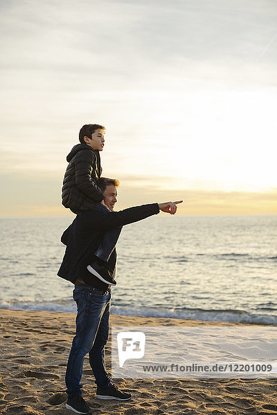 Father carrying son piggyback on the beach at sunset pointing finger