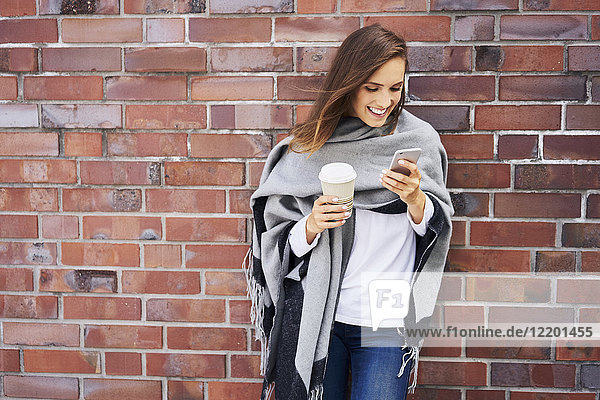 Happy woman with coffee to go looking at cell phone in front of brick wall