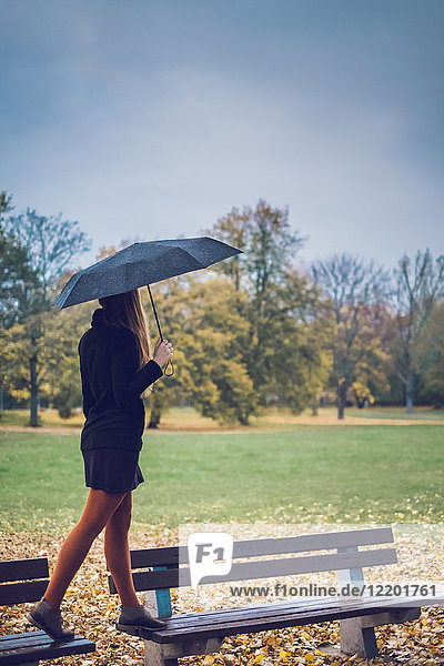 Young woman with umbrella balancing on two benches in autumnal park