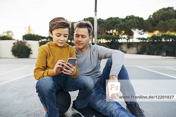 Father and son sitting on basketball outdoor court using cell phone