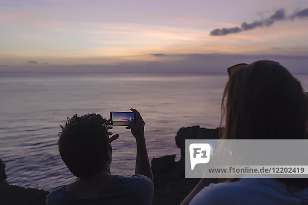 Indonesien  Bali  Lembonganische Insel  Freunde an der Meeresküste in der Abenddämmerung beim Fotografieren mit dem Handy