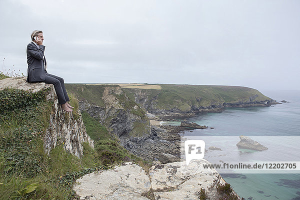UK  Cornwall  Gwithian  Geschäftsmann  der an der Küste sitzt und mit dem Handy telefoniert.