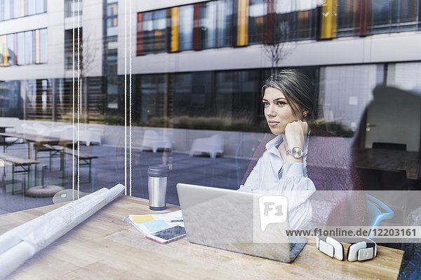 Young woman with laptop and documents behind windowpane