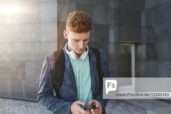 Rothaariger junger Mann im Freien mit Smartphone und Kopfhörer