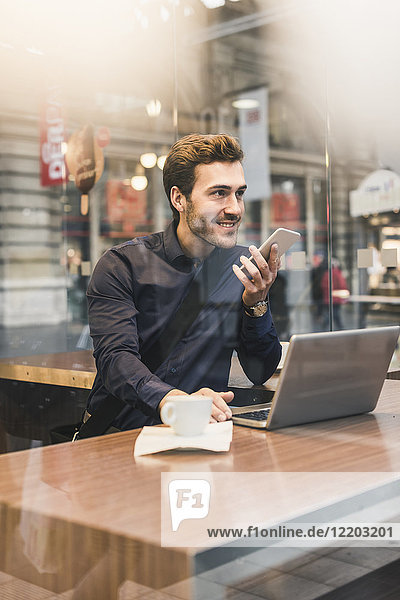 Lächelnder junger Geschäftsmann im Café am Bahnhof mit Handy und Laptop