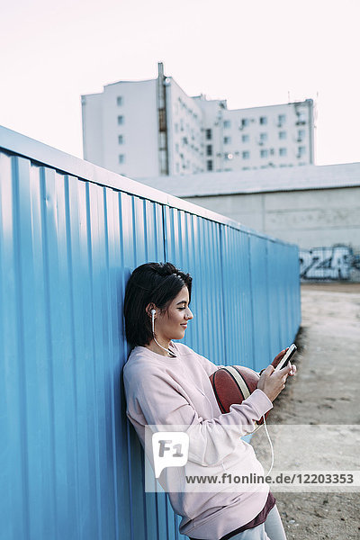 Junge Frau mit Basketball  Smartphone und Kopfhörer am Container