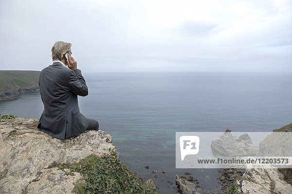 UK  Cornwall  Gwithian  Geschäftsmann  der an der Küste sitzt und mit dem Handy telefoniert.