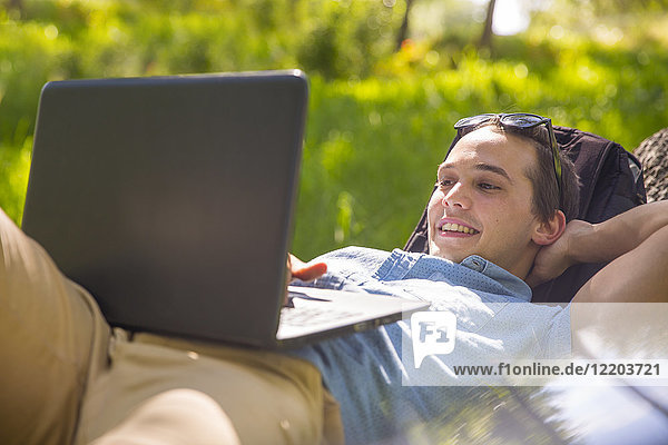 Portrait of happy young man lying on tree trunk in nature using laptop
