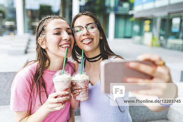 Two happy teenage girls drinking milkshakes in the city taking a selfie