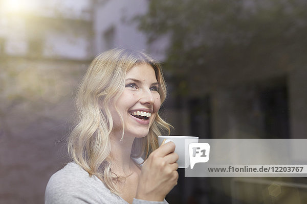 Porträt der lachenden Frau mit Tasse Kaffee hinter der Fensterscheibe