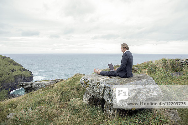 UK  Cornwall  Tintagel  businessman sitting on rock at the coast using laptop