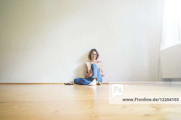 Lächelnde reife Frau sitzt auf dem Boden im leeren Raum mit Tablette