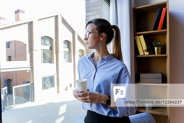Junge Frau mit Kaffeetasse mit Blick aus dem Bürofenster