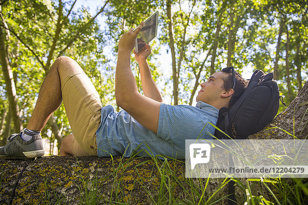 Mann liegt auf Baumstamm in der Natur und nimmt Selfie mit Tablette