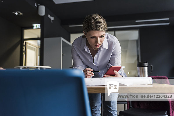 Junge Frau mit Handy und Plan am Tisch im Büro