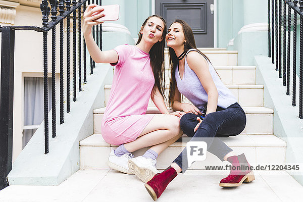Zwei fröhliche Teenager-Mädchen sitzen auf einer Treppe in der Stadt und nehmen einen Selfie.