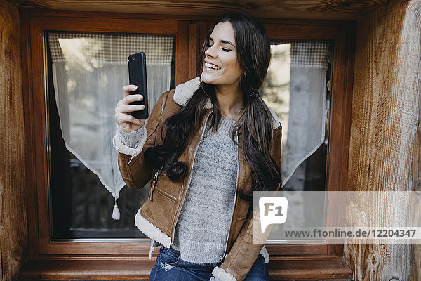 Fröhliche junge Frau am Fenster eines Holzhauses mit einem Selfie
