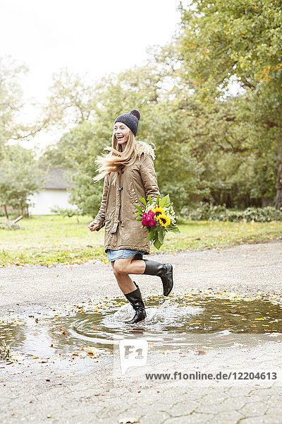 Happy young woman holding bunch of flowers jumping in puddle